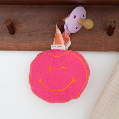 Handgemaakte roze en oranje smiley knuffel aan een geruit speenkoord, een kleurrijk en praktisch accessoire voor baby&